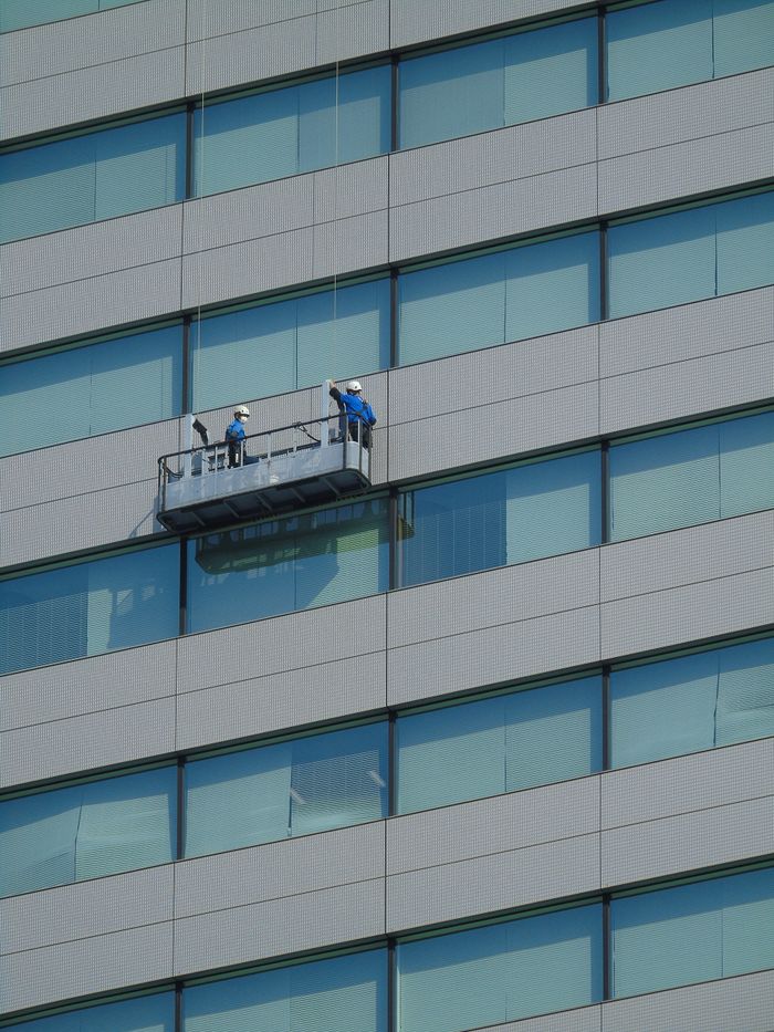 gallery-1 Two workers clean windows on a modern glass building facade using a suspended platform.
